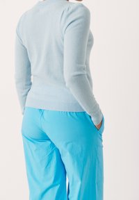 Persoon die een lichtblauwe trui met lange mouwen en felblauwe broek draagt, met handen in de zakken, gezien van achteren.