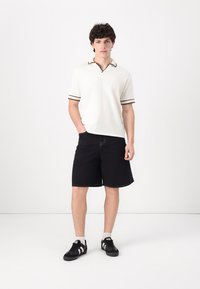 Vit kortärmad polo med V-hals och bruna accentränder; kombinerad med svarta shorts. Bärs med svarta sneakers med vita ränder.