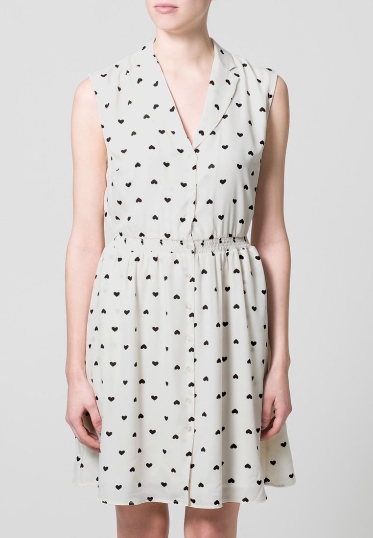 Robe sans manches en tissu crème clair avec un petit imprimé de cœurs noirs. Elle présente un devant boutonné et une taille élastique pour une silhouette ajustée.