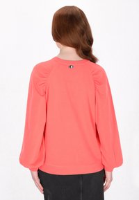 Pullover corallo con maniche lunghe a sbuffo, tessuto morbido, dettaglio arricciato sulle spalle, scollatura rotonda e un piccolo emblema decorativo sul retro.