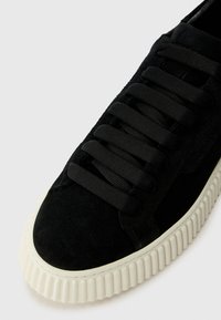 Black suede sneaker with thick off-white ribbed sole and wide black laces tied in a crisscross pattern on a white background.