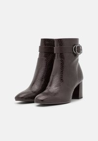Botas de cuero texturizado marrón oscuro hasta el tobillo con tacones en bloque y detalle de correa con una hebilla plateada sobre un fondo blanco.