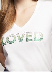 Bílý tričko s výstřihem do V, na kterém je slovo "LOVED" v gradientu od zelené po Tyrkysovou barvu s tečkovaným vzorem. Hladká textura látky.