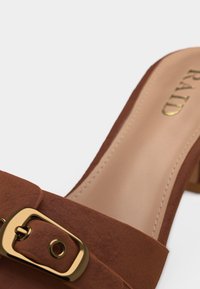 Gros plan sur une mule en daim marron avec une boucle dorée et une semelle intérieure beige marquée du nom "RALD".