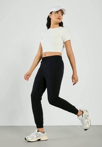 Zwarte slim-fit joggings met een trekkoord in de taille, gedragen met een cropped wit T-shirt en zilveren sportschoenen. Het model poseert tegen een neutrale achtergrond.