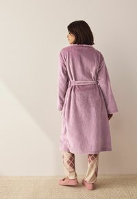 Lys lilla badekåbe med vaffelmønstret tekstur, lange ærmer og bindebælte, båret over creme- og lyserøde argyle-mønstrede bukser og lyserøde hjemmesko.