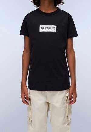 Osoba ubrana w czarny t-shirt Napapijri Geographic oraz jasne spodnie typu cargo, widoczna od szyi do bioder na jednolitym tle.