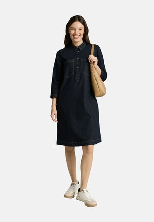 Femme souriante, portant une robe en jean foncé mi-longue avec col et boutons, des baskets blanches, et portant un sac à bandoulière en cuir marron clair.
