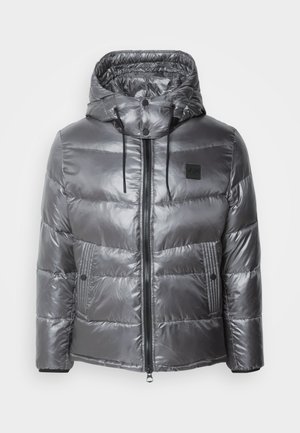 Chaqueta acolchada gris con un acabado brillante, capucha y cremallera frontal. Cuenta con dos bolsillos laterales y un parche negro con logo en el pecho.