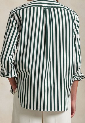 Persona que lleva una camisa blanca con rayas verticales verde oscuro y mangas remangadas, vista desde atrás.