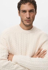 Maglione crema a trecce con scollo rotondo e polsini a coste, caratterizzato da un motivo testurizzato e una vestibilità ampia. Modello con le braccia incrociate.