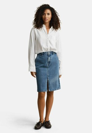 Femme debout portant une chemise blanche ample à boutons, une jupe en denim bleue longueur genou avec une fente à l'avant, et des chaussures bateau marron foncé.