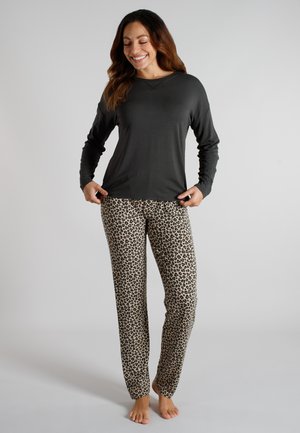 Woman smiling, wearing black long-sleeve shirt and beige leopard print pants, standing barefoot against a plain light background.