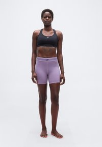 Αθλητικό άτομο όρθιο, ξυπόλητο, φοράει μαύρο αθλητικό μπουστάκι και μοβ στενά σορτς, μπροστά σε απλό φωτεινό φόντο.