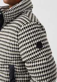 Zwart-witte houndstooth-patroonjas met hoge kraag en zwarte patch op de bovenarm, gedragen door een persoon.