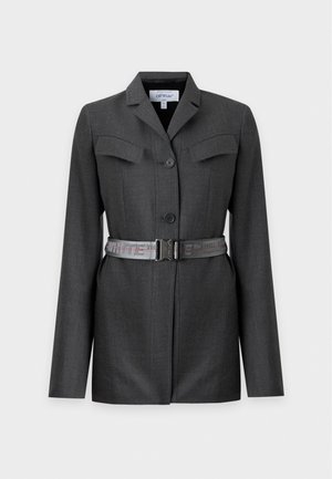 Mujer que lleva un vestido tipo blazer gris oscuro con bolsillos delanteros y cinturón, combinado con sandalias negras de tacón alto, de pie sobre un fondo liso.