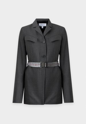 Mujer que lleva un vestido tipo blazer gris oscuro con bolsillos delanteros y cinturón, combinado con sandalias negras de tacón alto, de pie sobre un fondo liso.