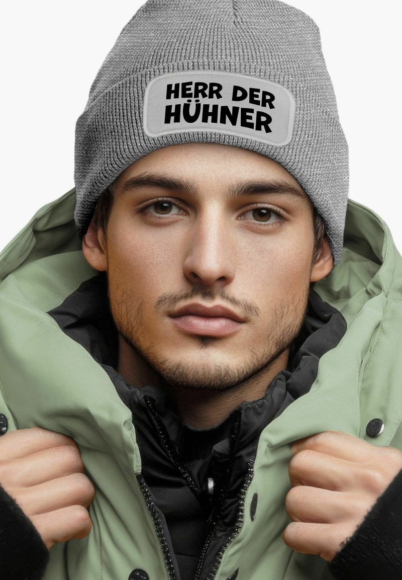 Gray knit beanie with a patch reading "HERR DER HÜHNER" on a flat crown, paired with a green and black puffer jacket. Textured materials visible.