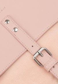 Rózsaszín műbőr pánt ezüst csattal és szegecsekkel, egy puha textúrájú felülethez rögzítve. Az emblémázott márkanevet tartalmaz.