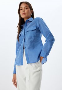 Camisa de botones a rayas azules con dos bolsillos en el pecho y mangas largas, con un dobladillo ligeramente curvado y una textura de tejido ligera.