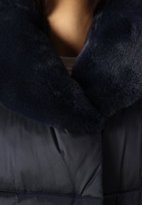 Mörkblå vadderad kappa med en mjuk, mörk pälskrage. Slätt yttertyg med quiltade detaljer synliga längs kroppen.