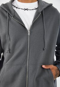 Felpa grigia con zip, dotata di un ampio cappuccio, tasche frontali e zip argentata. Il materiale sembra morbido con una texture liscia.