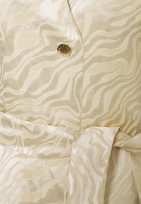 Veste en tissu beige tissé avec un motif de vagues subtil, dotée d'un bouton doré et d'une ceinture à nouer à la taille pour un détail supplémentaire.