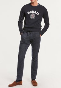 Homme portant un pull MORRIS bleu marine, un pantalon foncé à motifs tachetés avec cordon de serrage, et des mocassins en daim marron, debout les mains dans les poches.