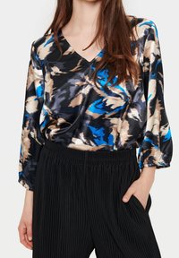 Satinblus med V-urt, med ett abstrakt mönster i svart, blått och beige. Matchas med svarta plisserade byxor.