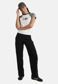 Short-sleeve white and green T-shirt with logo, paired with black wide-leg pants and black shoes, topped with a green cap.