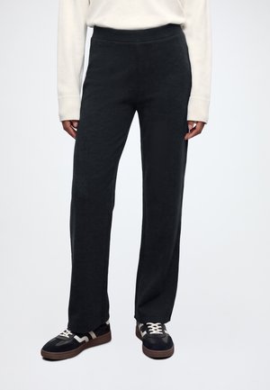 Persona que lleva pantalones negros de pierna recta, camiseta blanca de manga larga y zapatillas negras con cordones blancos, de pie frente a un fondo liso.