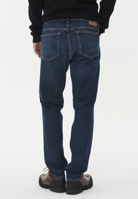 Vyras, vilkintis tamsiai mėlynus džinsus, rudos spalvos žygių batus ir juodą ilgomis rankovėmis marškinėlį, pavaizduotas iš nugaros prieš baltą foną.