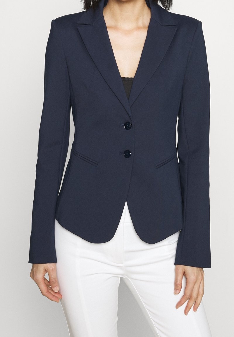 Granatowa marynarka o dopasowanym kroju, z dwoma przednimi guzikami, klapami z wycięciem i subtelnymi bocznymi kieszeniami, zestawiona z białymi dopasowanymi spodniami.