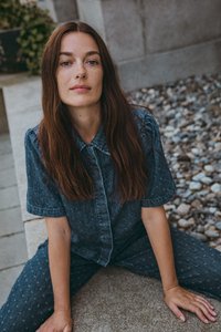 Donkerblauwe denim jumpsuit met korte mouwen, kraag en subtiele patronen. Model zit op een stenen oppervlak omringd door kiezels.