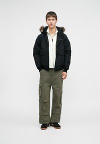 Μαύρο τζάκετ puffer με κουκούλα επενδεδυμένη με γούνα, δύο μπροστινές τσέπες, λευκή φλις φούτερ με φερμουάρ από μέσα, ελιά cargo παντελόνι και μαύρα αθλητικά παπούτσια.