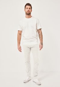 Weißes Baumwoll-T-Shirt mit kurzen Ärmeln, kombiniert mit hellen Jeans. Das Model trägt weiße Sneakers. Sichtbare Armtattoos.