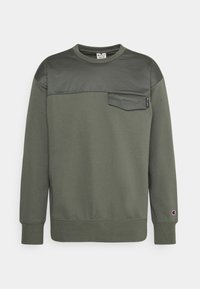 Felpa a manica lunga verde oliva con pannello superiore del torace di colore più scuro, piccolo taschino sul petto, polsini a coste e logo Champion sulla manica sinistra.