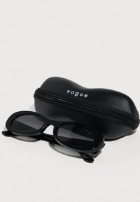 Μαύρα οβάλ γυαλιά ηλίου με σκούρα φακούς, ακουμπισμένα δίπλα σε μια μαύρη σκληρή θήκη με φερμουάρ με την ετικέτα "vogue." Ομαλή υφή πλαστικού σε όλη την επιφάνεια.