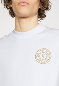 T-shirt bianco a girocollo in cotone con una texture morbida. Presenta un logo circolare ricamato in oro che include "Versace Jeans Couture."
