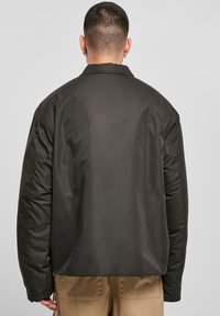 Giacca nera imbottita con colletto alto e vestibilità rilassata, caratterizzata da una texture liscia e una superficie leggermente lucida. Nessun hardware visibile.