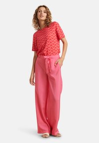 Rotes, gemustertes Kurzarmoberteil mit geometrischen Designs, kombiniert mit lockeren rosa Hosen mit Bindegürtel. Glatter Stoff und entspannte Passform.