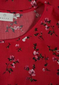 Tissu floral rouge avec petites fleurs roses et blanches et feuilles vertes, montrant l'encolure et l'étiquette du vêtement indiquant la taille 38.