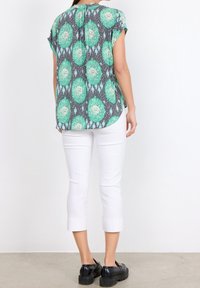 Blusa a maniche corte in tessuto leggero con base grigia, caratterizzata da motivi circolari verdi. Abbinata a pantaloni bianchi a tre quarti e scarpe nere.
