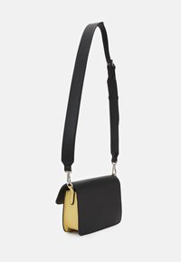 Schwarze Ledereinkaufstasche mit gelbem Innenfutter, mit Klappenverschluss und einem verstellbaren, schwarzen Riemen mit metallischen Beschlägen.