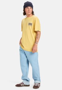 Muž oblečený v žlutém grafickém tričku, světlých modrých džínách, hnědých teniskách s bílými pruhy a tmavě modré čepici, stojící a dívající se do strany.