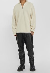 Osoba v béžovém dlouhém pulovru s půlzipem, černých kožených kalhotách, černých robustních botách a vrstvených náhrdelnících.