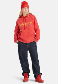 Sudadera roja con el logotipo amarillo de "Timberland", combinada con jeans holgados de color azul oscuro y botas rojas, rematada con un gorro rojo a juego.