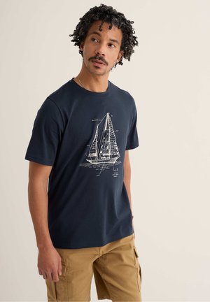 Mężczyzna z kręconymi włosami, ubrany w granatowy T-shirt z białym diagramem żaglówki i beżowe krótkie spodnie typu cargo, patrzący nieco na bok.