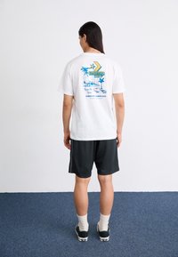 Hvit t-skjorte med fargerik "CONVERSE MOTEL" grafikk på baksiden, kombinert med svarte shorts, hvite sokker og mørke sko på blå teppe.