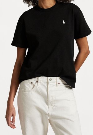 Persoon draagt een zwart T-shirt met korte mouwen met een klein wit Polo-logo en witte jeans met hoge taille en voorzakken.